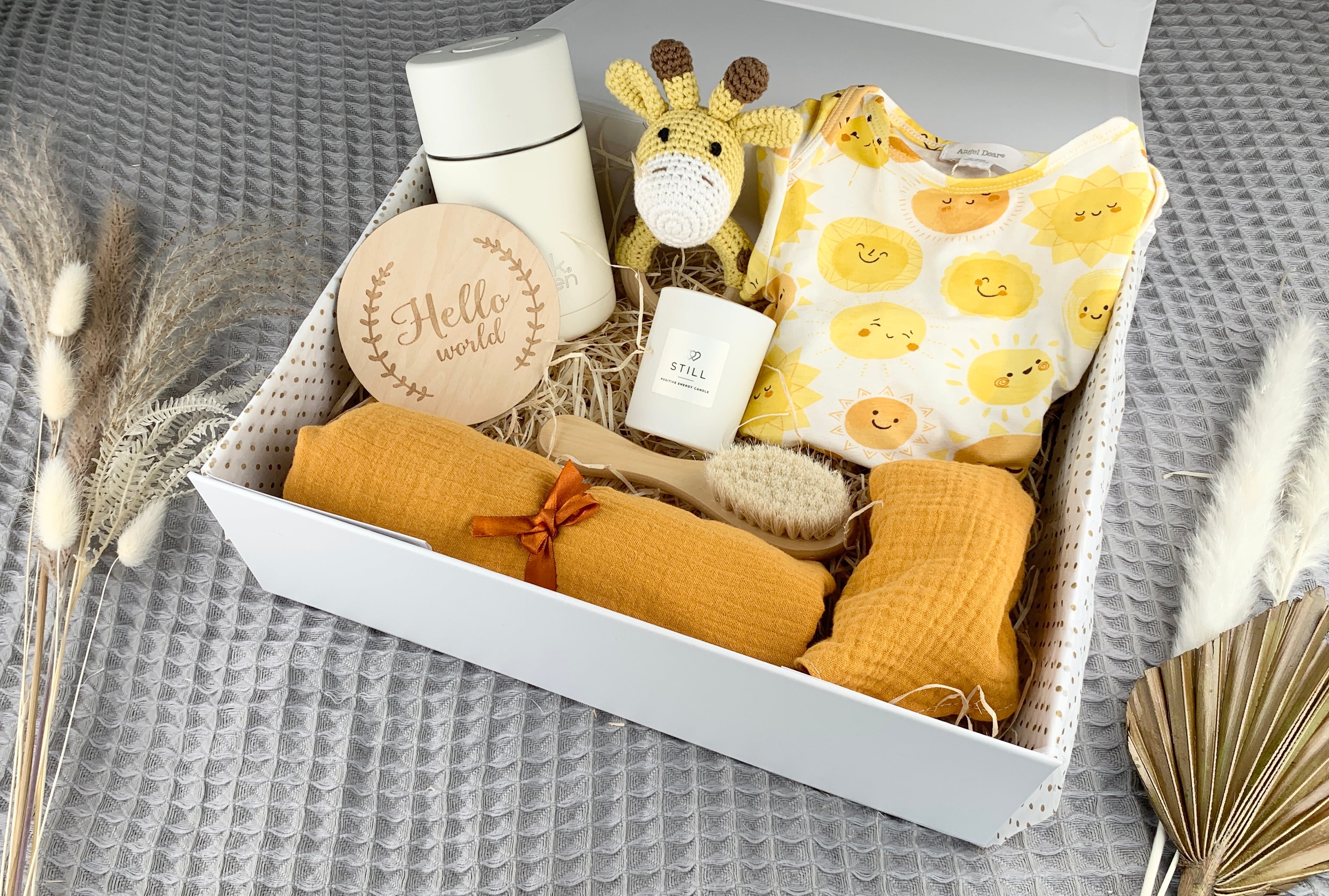 Large sunshine baby gift box containing yellow crochet giraffe rattle, Angel Dear sunshine baby grow, wooden brush, 100% cotton mustard bib and muslin, Still candle, Frank Green ceramic coffee cup and Hello world wooden announcement disc